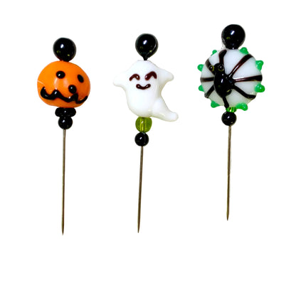 Three Halloween-themed glass bead pins on metal needles, featuring an orange jack-o'-lantern, a white smiling ghost, and a black spider with green spikes, displayed on a textured white surface.
[additional_image_link]
