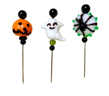 Three Halloween-themed glass lampwork beads on metal pins, featuring an orange jack-o'-lantern, a white ghost, and a black spider with green accents, displayed on a white background.
[image_link]