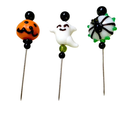 Three Halloween-themed glass pinback beads on metal pins, featuring an orange pumpkin with black facial features, a white ghost with a black outline, and a black-and-white spider with green accents, displayed on a textured white surface.
[additional_image_link]