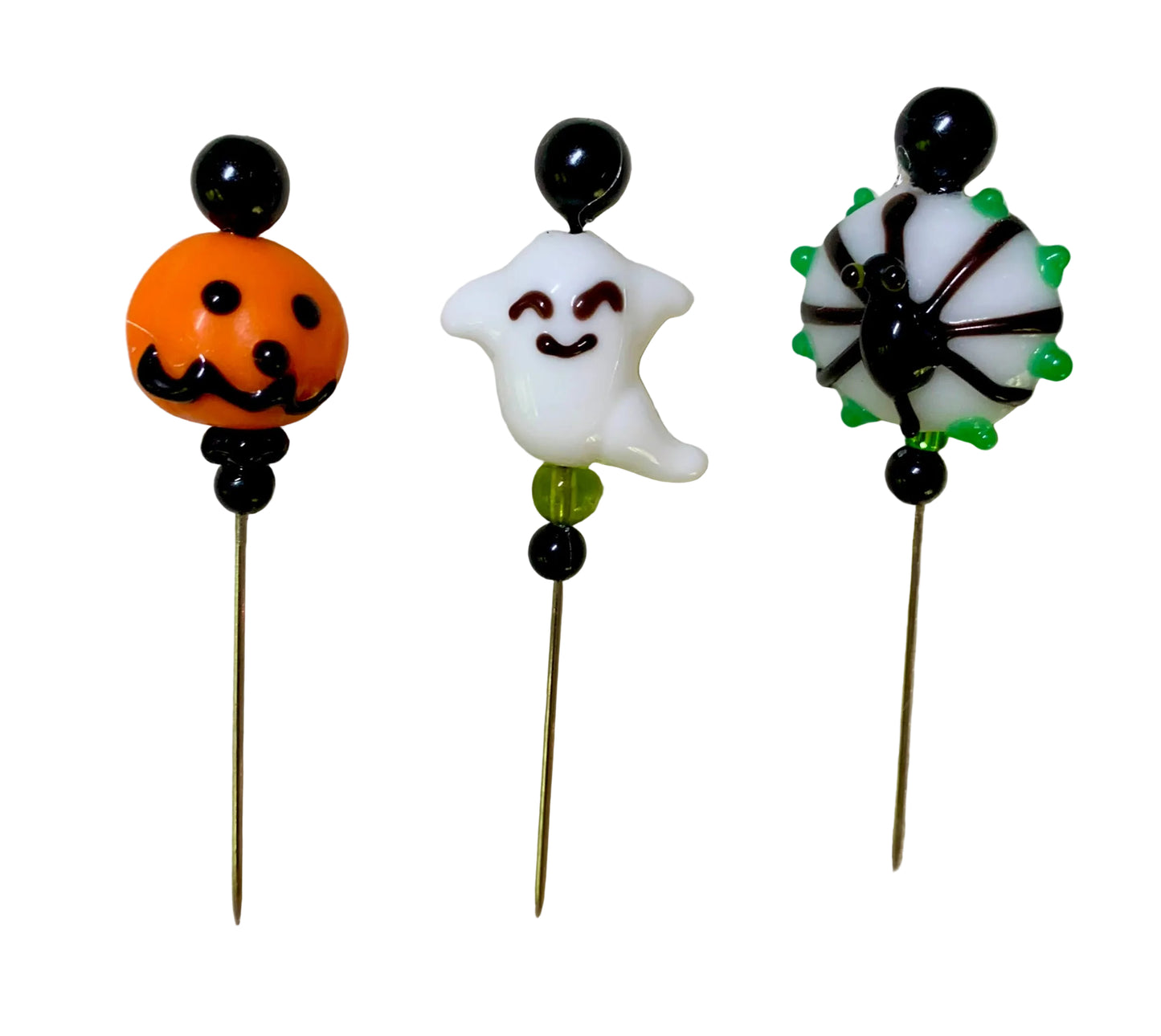 Three Halloween-themed glass charms on thin metal pins, featuring an orange jack-o'-lantern, a white smiling ghost, and a black spider on a white background with green accents, displayed on a white surface.
[additional_image_link]