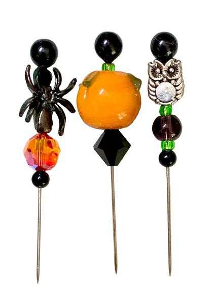 Three Halloween-themed beaded pins on metal needles, featuring a black spider, an orange pumpkin with green leaves, and a silver owl, set against a white background.
[image_link]