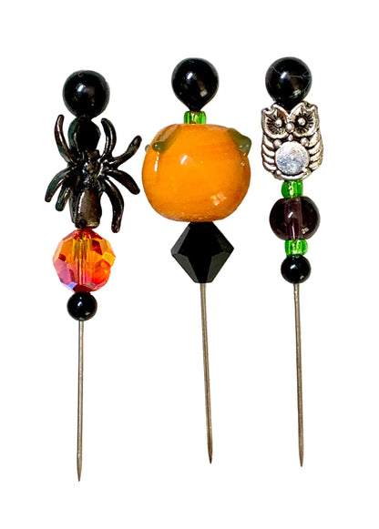 Three Halloween-themed beaded pins displayed on a white background. The left pin features a black spider, a faceted orange and red bead, and black beads. The right pin is a silver owl. The middle pin has a lampwork pumpkin.
[additional_image_link]
