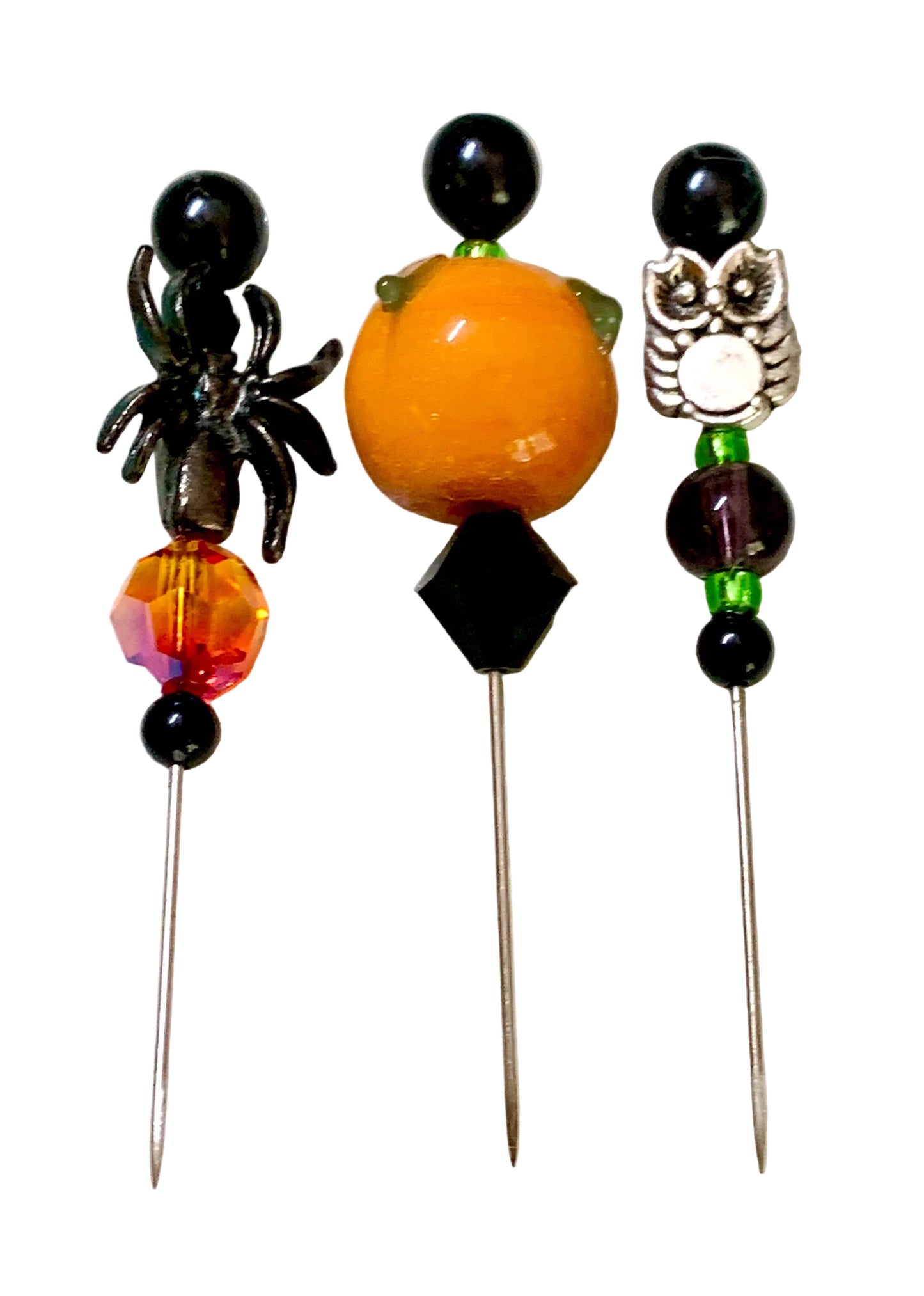 Three decorative beaded pins with metal pins, featuring a black spider and a silver owl charm,  an orange lampwork pumpkin with green accents, displayed on a white surface.
[additional_image_link]