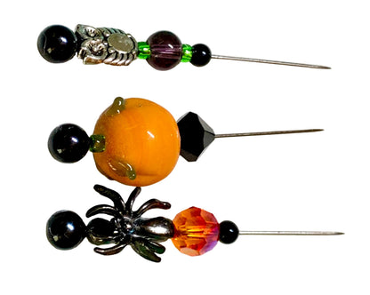 Three Halloween-themed beaded pins displayed on a white background. The left pin features a black spider, a faceted orange crystal, and a black bead. The right pin features a silver owl with purple and green beads. The middle bead is lampwork pumpkin with a black bicone.
[additional_image_link]
