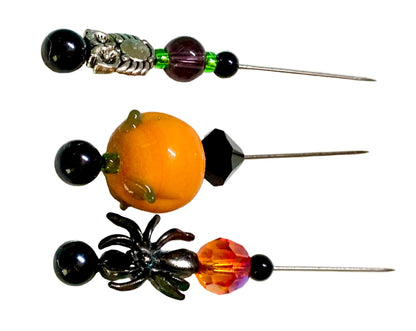 Three Halloween-themed beaded pins on a white background. The left pin features a black spider, a faceted orange and pink crystal bead, and black beads. The right pin a silver owl with green and purple beads. The middle pin is lampwork pumpkin with a black bicone.
[additional_image_link]