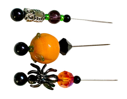 Three Halloween-themed beaded pins displayed on a white background. The left pin features a black spider and a faceted orange crystal. The right pin a silver owl with green and purple beads. The middle pin is lampwork pumpkin with a black bicone.
[additional_image_link]