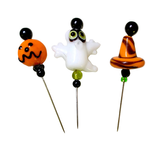 Three Halloween-themed glass bead pins on metal needles, featuring an orange jack-o'-lantern, a white ghost with green eyes, and an orange witch hat, displayed on a white background.
[image_link]