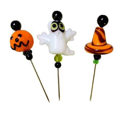 Three Halloween-themed glass pins on a white background: an orange jack-o'-lantern with black facial features and black bead accents, a white ghost with green eyes, and an orange-and-black witch hat with green bead accents.
[additional_image_link]