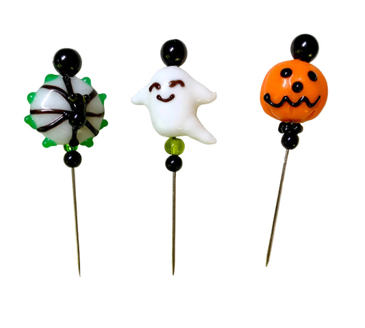 Three Halloween-themed glass lampwork pins displayed on a white background. The left pin features a white spider with black webbing and green accents on a black stem. The middle pin has a smiling ghost. The right pin has a jack-o'-lantern.
[image_link]
