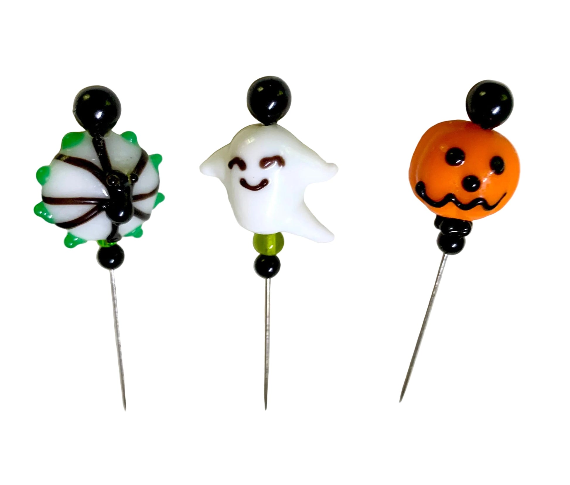 Three Halloween-themed glass bead pins on a white textured background. The right pin features an orange jack-o'-lantern with black details, the middle pin features a white smiling ghost with a black face, and the left pin features a white spider with green and black details.
[additional_image_link]
