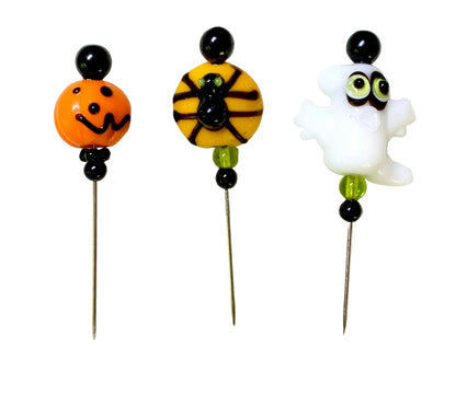 Three Halloween-themed beaded pins on a white background. The left pin features an orange jack-o'-lantern bead with a black face, the middle pin has a yellow and black spider  on a bead, and the right pin shows a white ghost bead with green eyes.
[image_link]