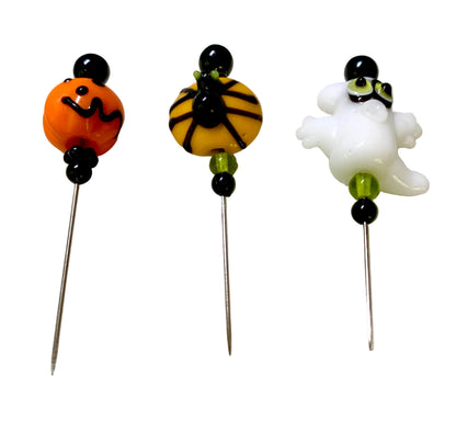 Three Halloween-themed glass bead pins on metal needles, featuring an orange jack-o'-lantern, a spider on a yellow bead, and a white ghost, displayed on a white surface.
[additional_image_link]