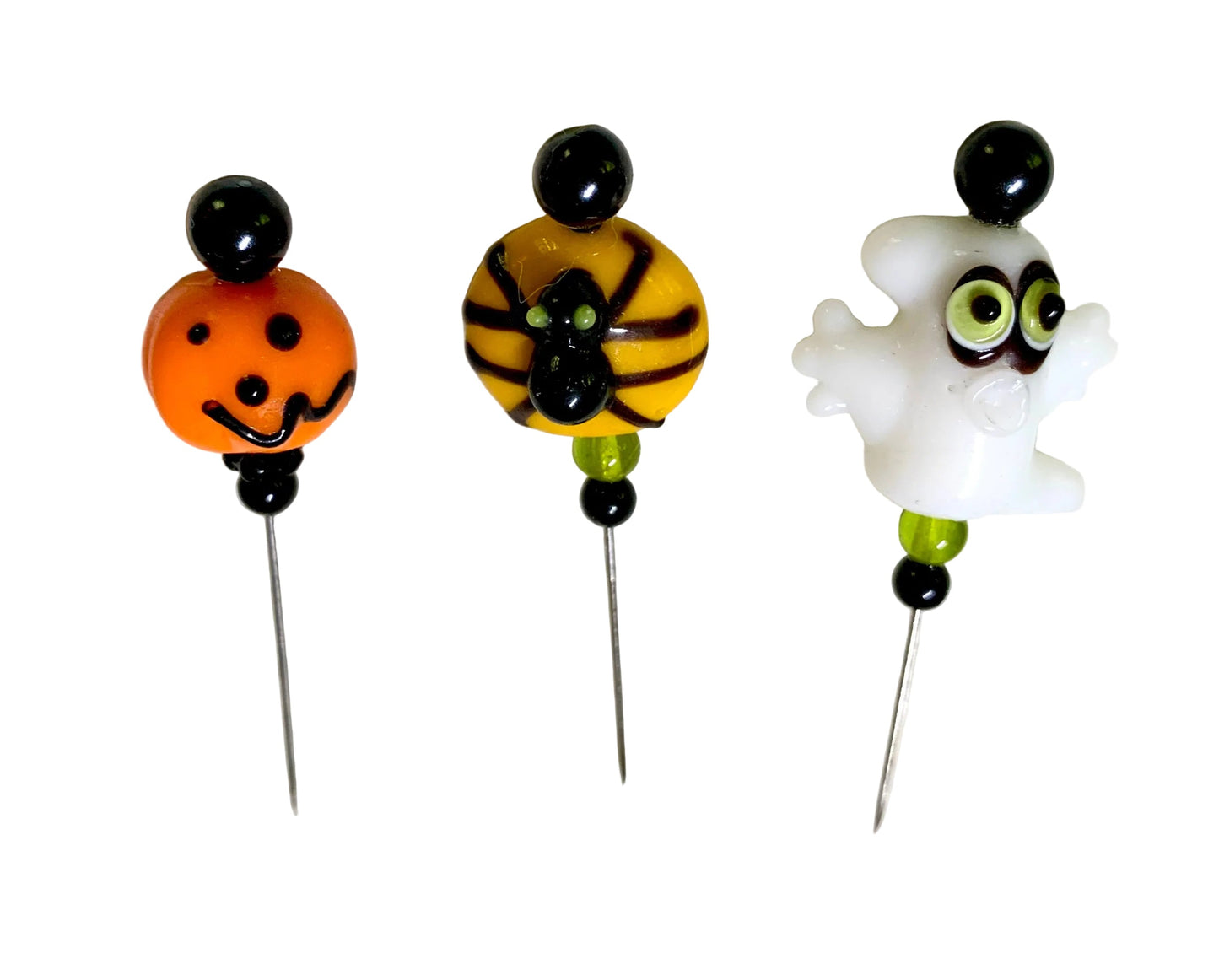Three Halloween-themed beaded pins displayed on a white surface. The pins feature a white ghost with green eyes, a yellow and black spider, and an orange jack-o'-lantern, with black and green beads.
[additional_image_link]
