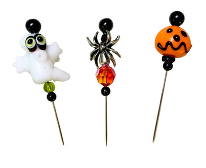 Three Halloween-themed glass beads on metal pins, featuring a white ghost with green eyes, a black spider with an orange and red faceted bead, and an orange jack-o'-lantern face.
[additional_image_link]