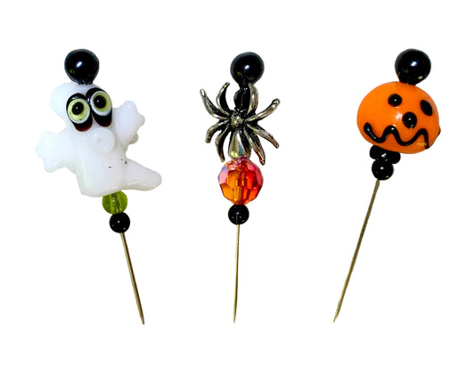 Three Halloween-themed glass beads on metal pins, featuring a white ghost with green eyes, a black spider with an orange and red faceted bead, and an orange jack-o'-lantern face.
[image_link]