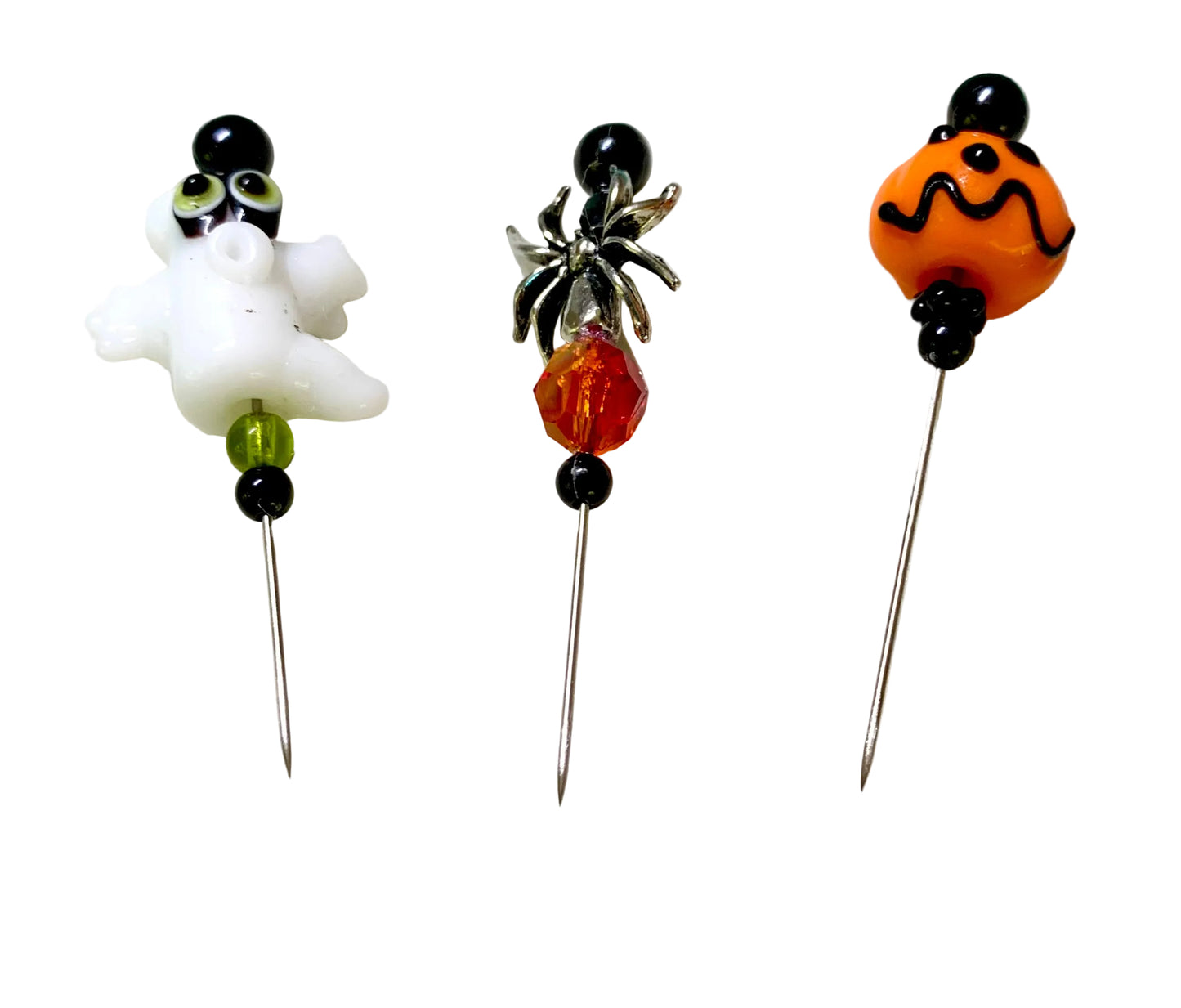 Three Halloween-themed beaded sewing pins displayed on a white surface. The left pin features a white ghost with green eyes and black beads. The right bead is a jack-o'-lantern. The middle bead is a silver spider. There are multiple complimentary beads in green, black and fire red.
[additional_image_link]