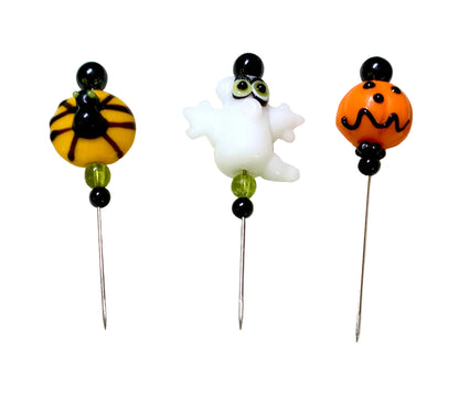 Three Halloween-themed glass beaded pins on a white background. One pin features a spider on a yellow bead, one pin features a white ghost, and one pin features an orange jack-o'-lantern.
[additional_image_link]