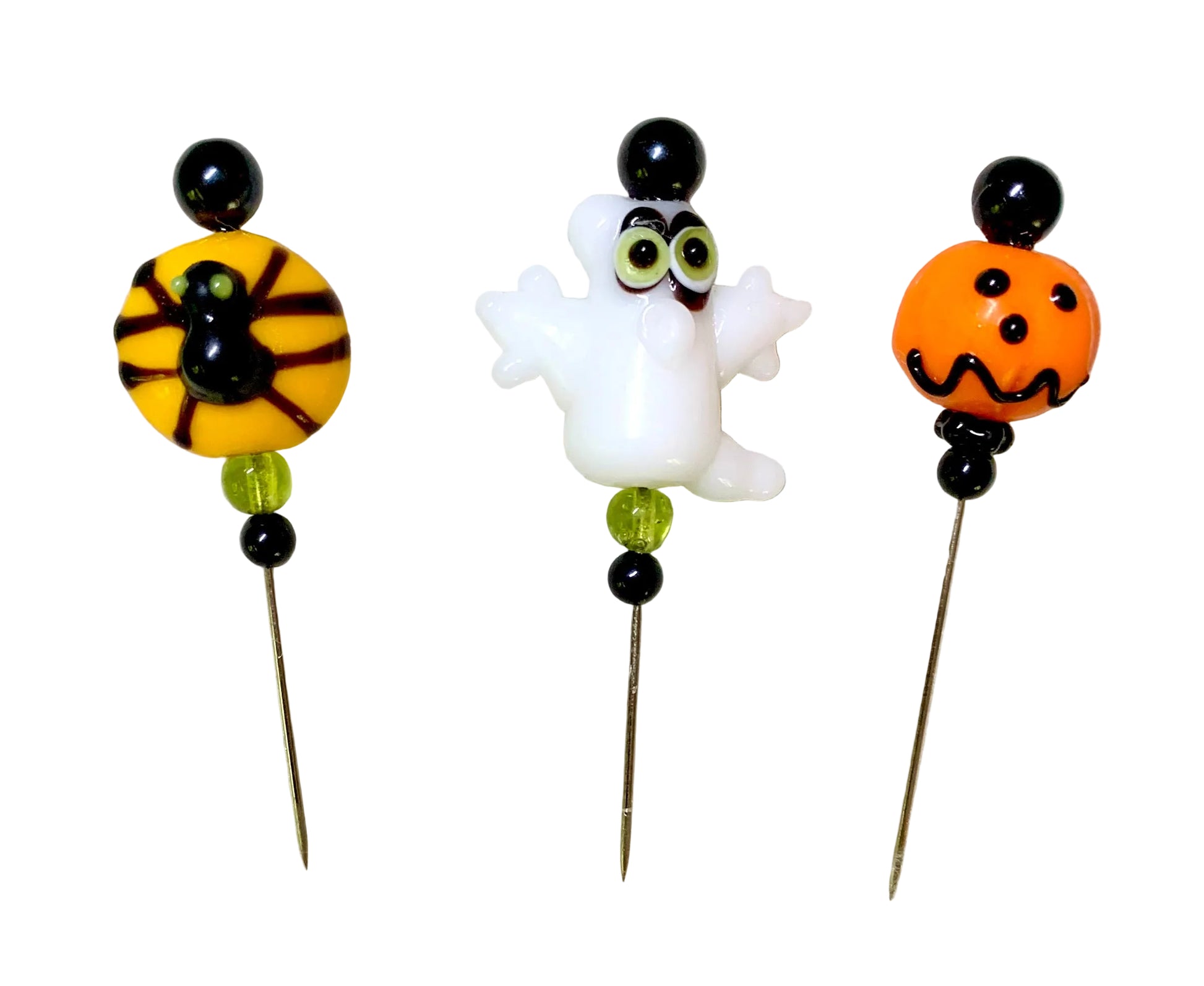 Three Halloween-themed glass  beaded pins on a white background. One pin features a spider on a yellow bead, one pin features a white ghost, and one pin features an orange jack-o'-lantern.
[additional_image_link]