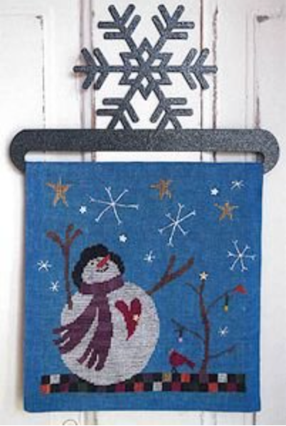 A square blue cross stitch featuring a winter snowman design, hung on a white wooden door with a dark metal snowflake ornament above it.
Photo by SamSarah Studio Designs [image_link]