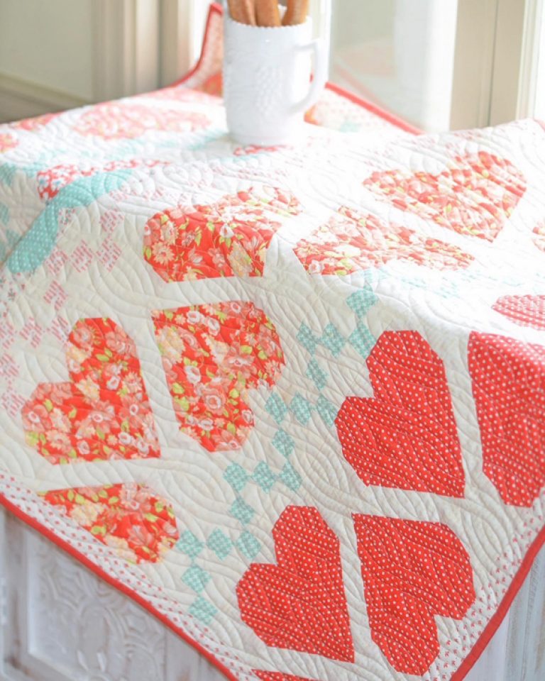 A quilt 'Fair Isle Hearts' by Joanna Figueroa, showing a quilt with red and white heart shapes on a white background, and a detail of a quilt block with various red prints.
Photo by Fig Tree & Co. [additional_image_link]