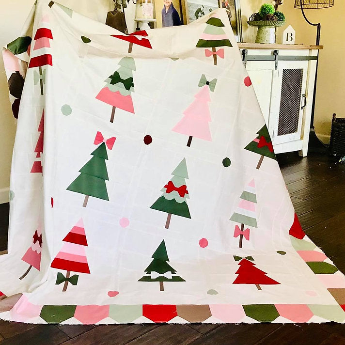 A white fabric throw blanket featuring a playful pattern of Christmas trees in green, pink, and red, with geometric border shapes, draped over a chair in a home setting.
Photo by My Sew Quilty Life [additional_image_link]