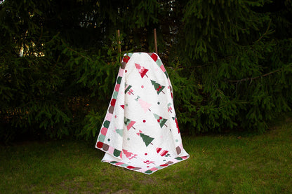 A white quilted blanket with a Christmas tree and dot pattern in red, green, and pink, hanging outdoors on a clothesline against a backdrop of green trees and grass.
Photo by My Sew Quilty Life [additional_image_link]