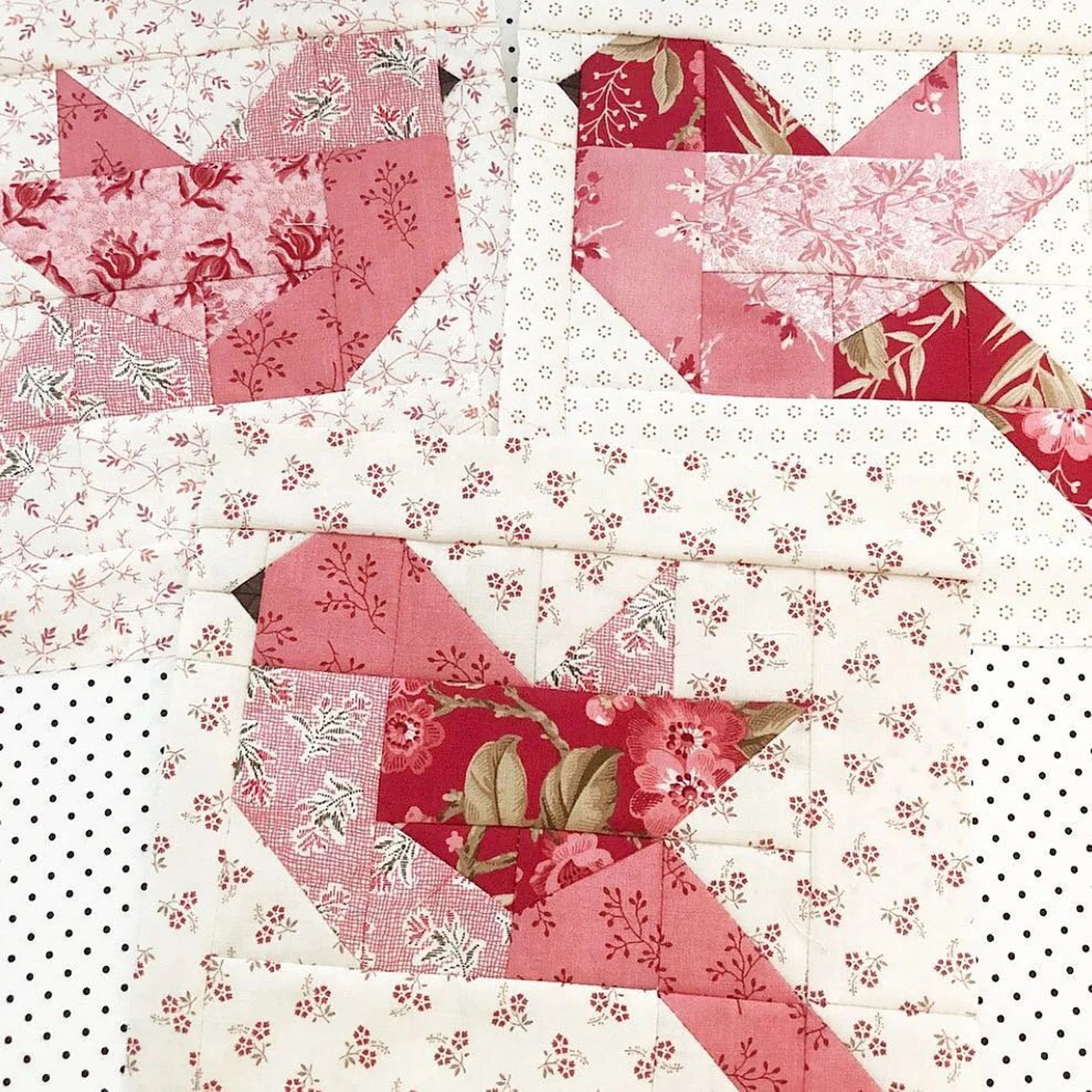 A close-up of a quilted textile featuring three stylized bird shapes stitched in pink, red, and floral patterned fabrics over an off-white dotted background.
Photo by ThePatternBasket.com [additional_image_link]