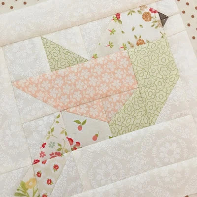 A close-up of a quilted fabric panel featuring a geometric bird design made from patterned fabric pieces in soft peach, sage green, and floral prints on an off-white background.
Photo by ThePatternBasket.com [additional_image_link]