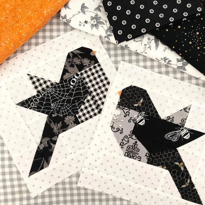 Two Halloween-themed bird quilt blocks displayed on a checkered fabric background, surrounded by other patterned fabrics including orange glitter, black and white polka dots, and black with white spiderwebs and honeycomb designs.
Photo by ThePatternBasket.com [additional_image_link]