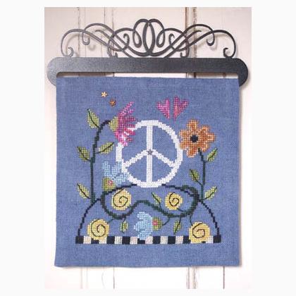 A blue cross-stitch sampler featuring a white peace sign surrounded by colorful flowers and green leaves, hung on a black metal wall hanger against a white wood paneled wall.
Photo by SamSarah Studio Designs [additional_image_link]