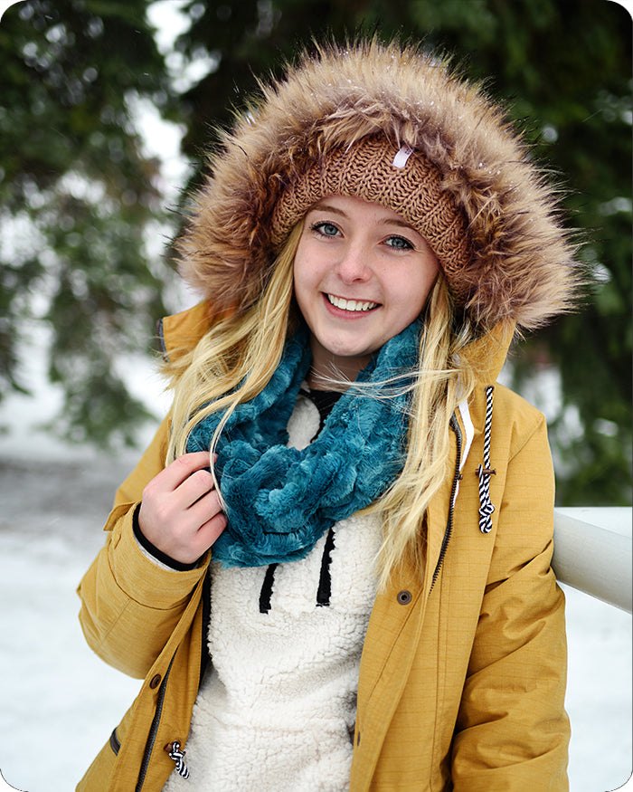 A smiling young woman outdoors in the snow wearing a mustard yellow winter parka with a brown faux-fur trimmed hood, a brown knit beanie, and a chunky teal blue scarf.
Photo by Shannon Fabrics [additional_image_link]
