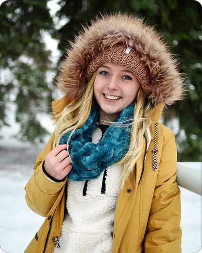 A smiling young woman outdoors in the snow wearing a mustard yellow winter parka with a brown faux-fur trimmed hood, a brown knit beanie, and a chunky teal blue scarf.
Photo by Shannon Fabrics [additional_image_link]