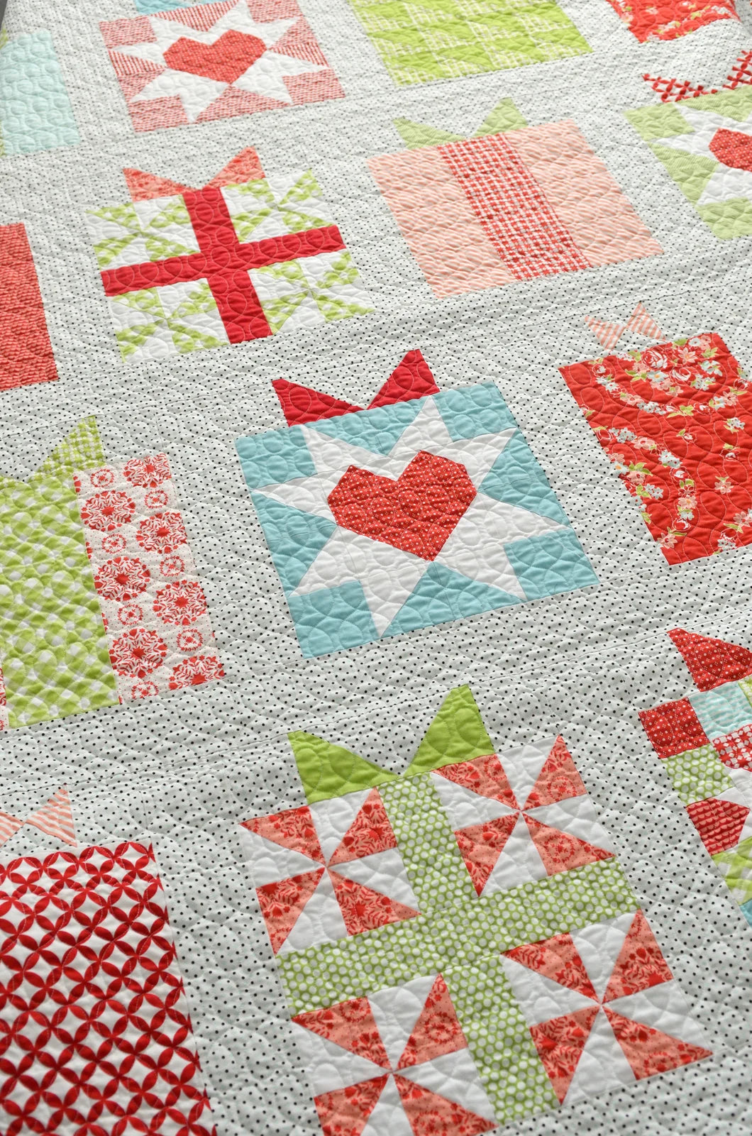 A close-up of a quilted patchwork blanket featuring various gift-themed blocks in red, green, pink, and light blue on a white textured background.
Photo by Thimble Blossoms [additional_image_link]