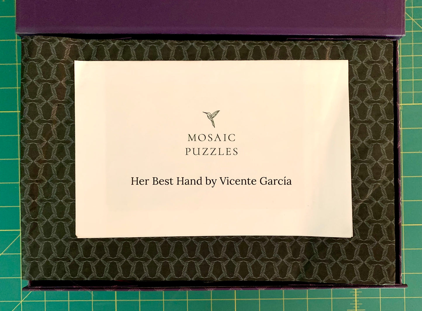 A boxed puzzle product shown from above on a green cutting mat. The top of the box is purple, and the inner lid has a dark olive-green patterned surface.
Photo by Karen King [additional_image_link]