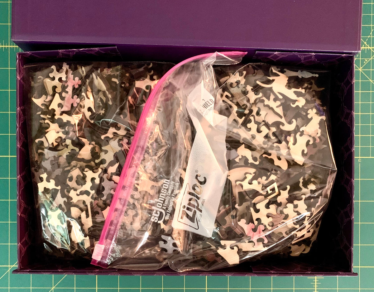 A purple box containing two clear plastic zip bags filled with jigsaw puzzle pieces in a black, gray, and beige camouflage pattern, placed on a green cutting mat.
Photo by Karen King [additional_image_link]