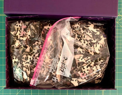 A purple box containing two clear plastic zip bags filled with jigsaw puzzle pieces in a black, gray, and beige camouflage pattern, placed on a green cutting mat.
Photo by Karen King [additional_image_link]