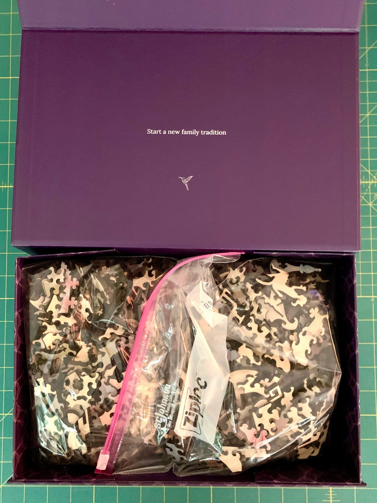 An open purple gift box containing a camouflage-patterned garment in a clear zip bag, placed on a green cutting mat. The inside lid has white text reading 'Start a new family tradition' with a small logo below it.
Photo by Karen King [additional_image_link]