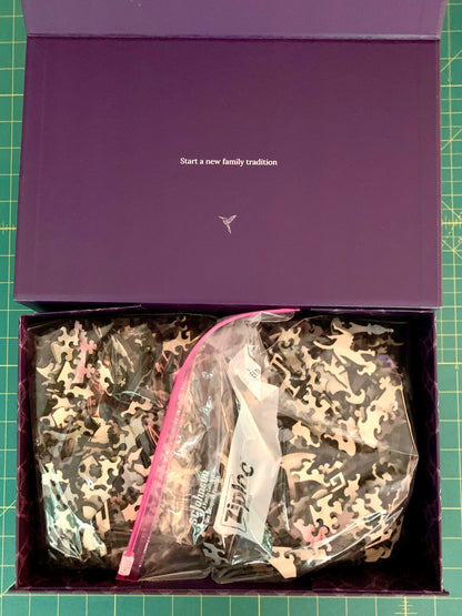 An open purple gift box containing a camouflage-patterned garment in a clear zip bag, placed on a green cutting mat. The inside lid has white text reading 'Start a new family tradition' with a small logo below it.
Photo by Karen King [additional_image_link]