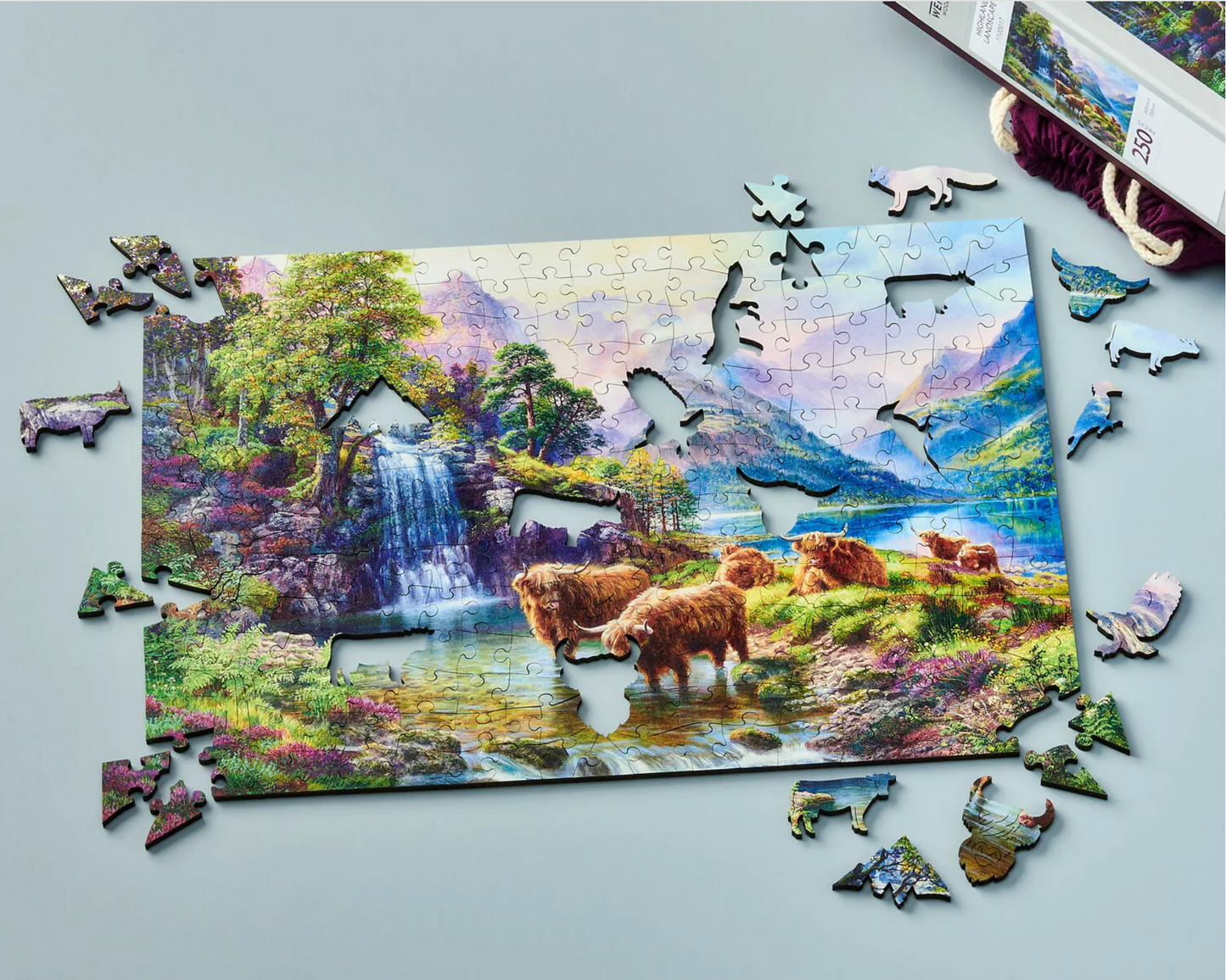 A wooden jigsaw puzzle depicting a highland landscape scene with cows, a waterfall, and mountains in the background.
Photo by Wentworth Puzzles [additional_image_link]