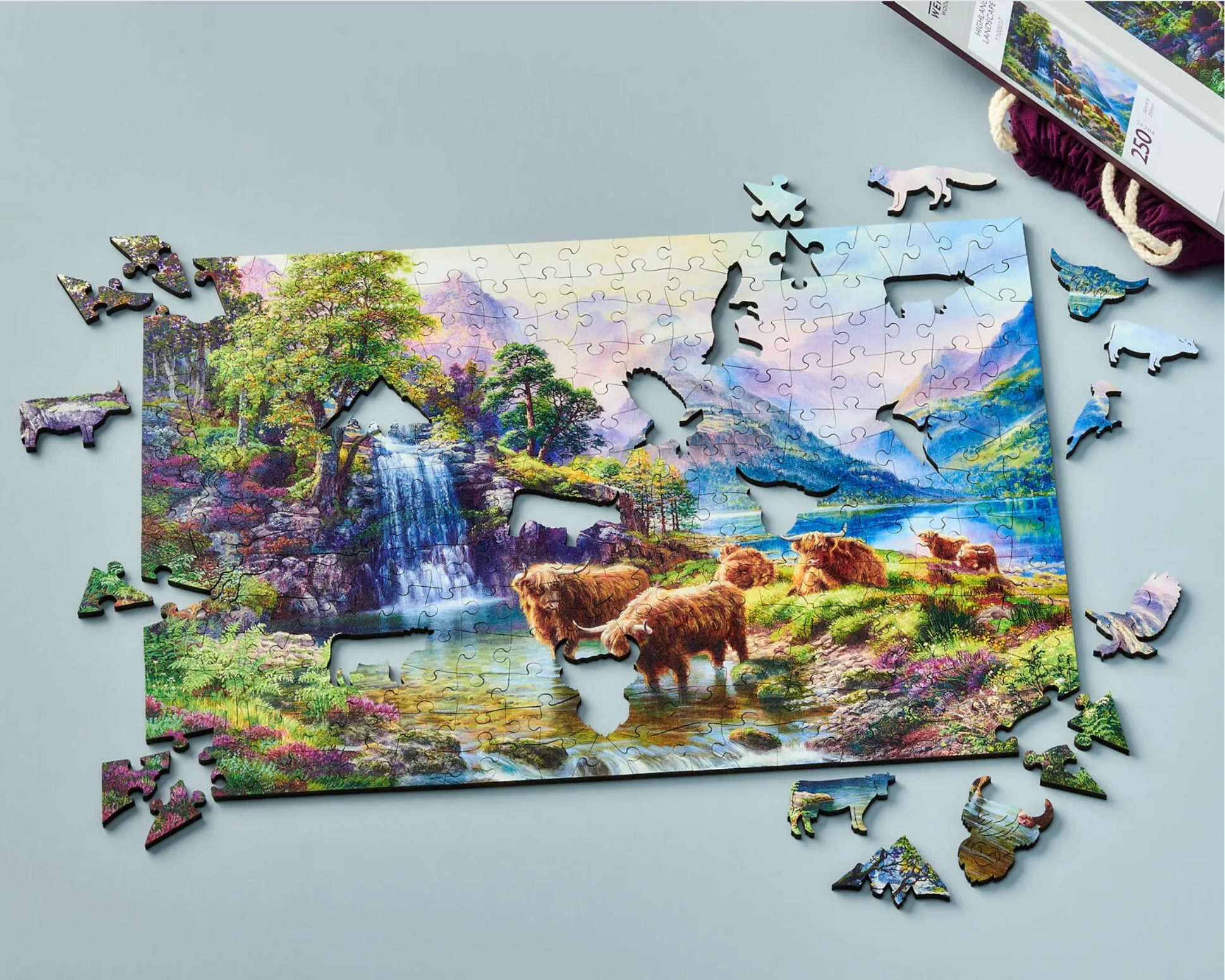A wooden jigsaw puzzle depicting a highland landscape scene with cows, a waterfall, and mountains in the background.
Photo by Wentworth Puzzles [additional_image_link]