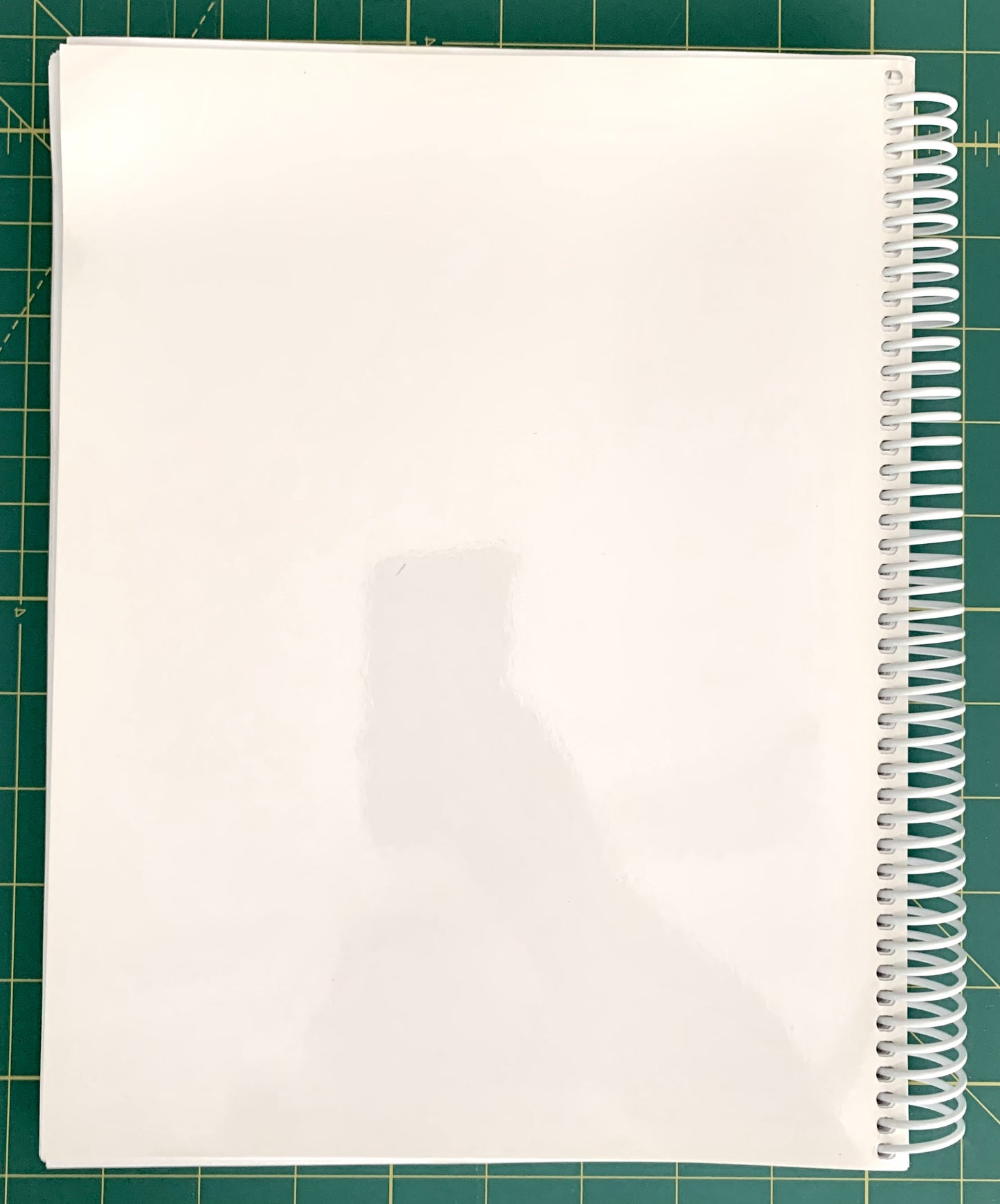 A white spiral-bound notebook with a glossy cover, photographed from above on a green cutting mat with grid lines.
[additional_image_link]