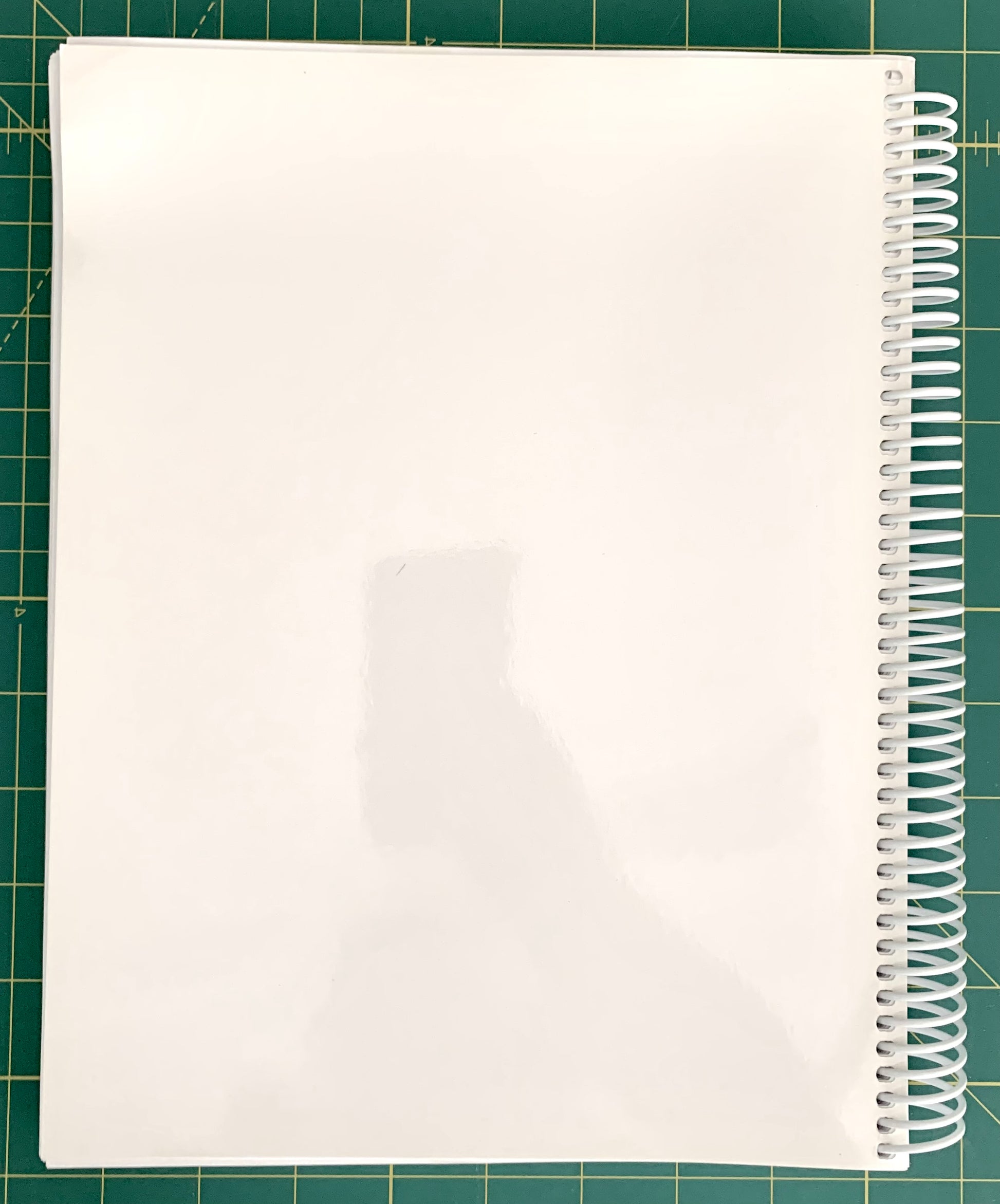 A white spiral-bound notebook with a glossy cover, photographed from above on a green cutting mat with grid lines.
[additional_image_link]