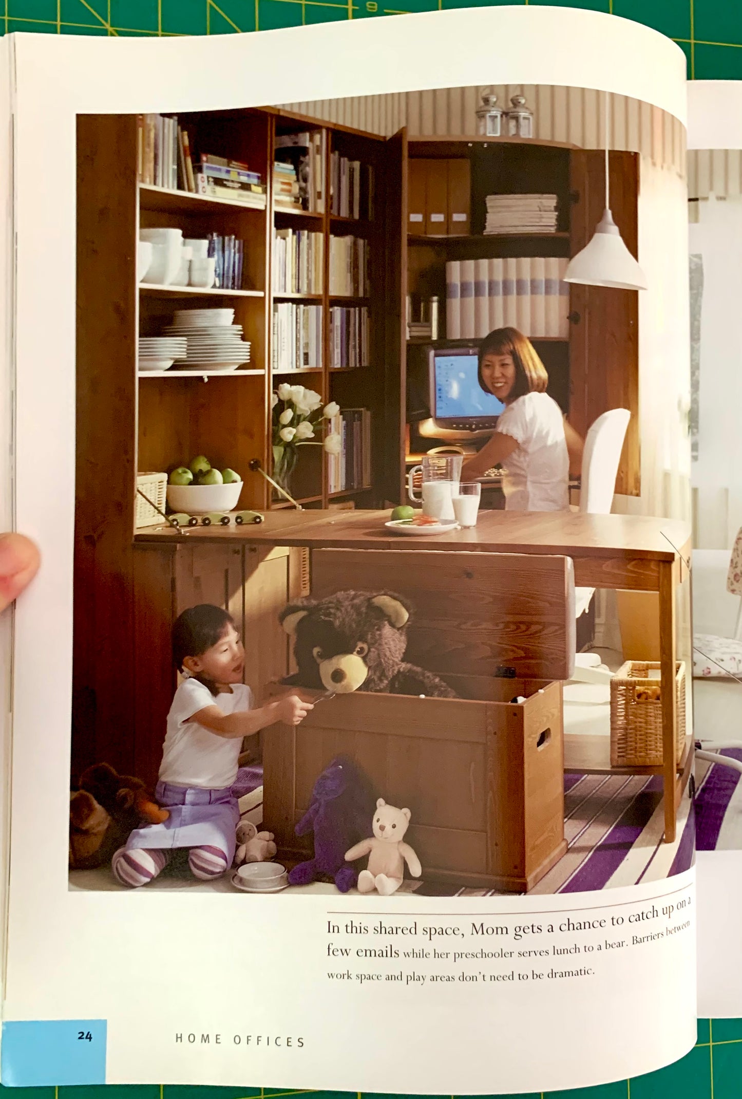 A printed interior design magazine page showing a home office layout with shelving, desk, computer, and children playing with stuffed animals in a playpen setup. The page includes a photo of the space and a block of text at the bottom.
[additional_image_link]