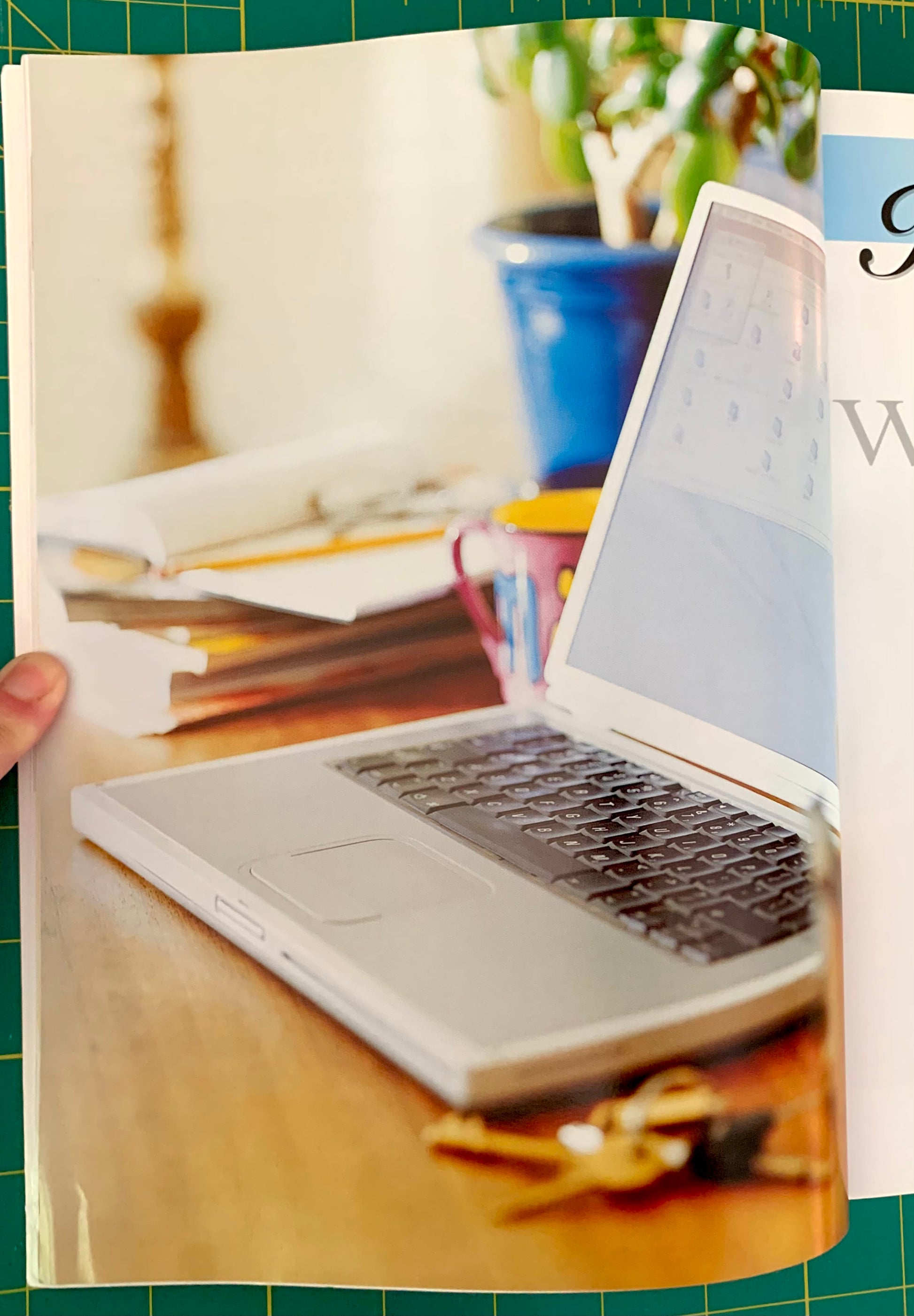 A printed page or booklet showing a laptop on a wooden desk with papers, a mug, and keys, photographed against a green cutting mat. A hand is visible holding the left edge of the page.
[additional_image_link]