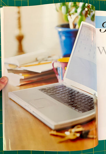 A printed page or booklet showing a laptop on a wooden desk with papers, a mug, and keys, photographed against a green cutting mat. A hand is visible holding the left edge of the page.
[additional_image_link]