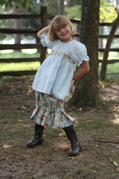 A young girl with blonde hair stands outdoors in a wooded area wearing a light blue smocked blouse, a floral print skirt, and dark brown boots.
Photo by Olabelhe [additional_image_link]