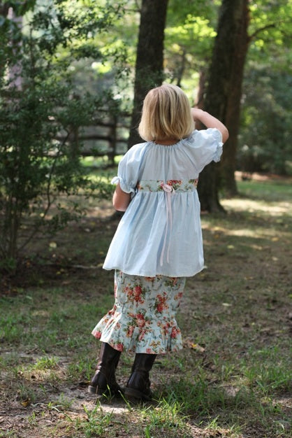 A young girl seen from behind in an outdoor wooded setting, wearing a light blue short-sleeve dress with a floral skirt and dark boots.
Photo by Olabelhe [additional_image_link]