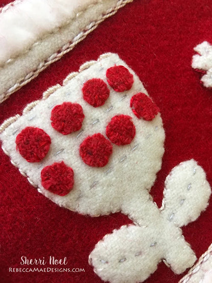 Close-up of a handmade felt Christmas ornament or decorative applique featuring a white holly leaf with red felt berries on a red felt background.
Photo by Rebecca Mae Designs [additional_image_link]
