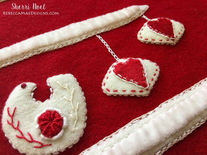 Handmade felt holiday ornaments on a red felt background, including a white goose with a red flower and two white heart ornaments with red hearts, with cream-colored braided trim.
Photo by Rebecca Mae Designs [additional_image_link]