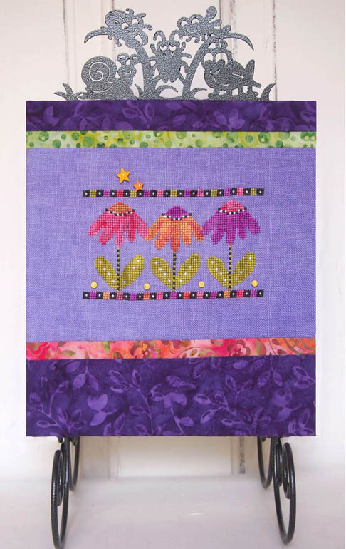 A square fabric art piece or quilt panel with a purple background and embroidered floral motifs in pink, orange, and yellow, framed by patterned purple borders and displayed on a black metal stand.
Photo by SamSarah Studio Designs [image_link]